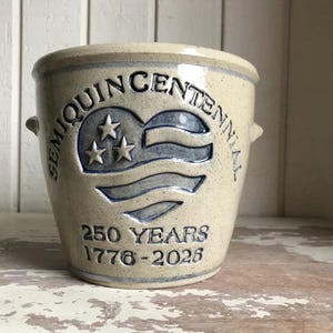 May include: A beige ceramic pot with blue lettering and a blue flag design. The text reads "SEMIQUINCENTENNIAL" and "250 YEARS 1776-2026". The pot has two small handles and a slightly flared rim. The design is a patriotic theme.