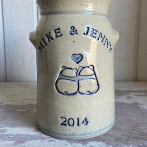 Può includere: Un vaso in ceramica beige con lettere blu e un design a forma di cuore blu. Il vaso dice "Mike & Jenny 2014".