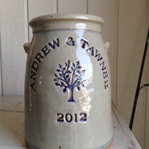 May include: A grey ceramic jar with blue lettering that reads "Andrew & Tawnie 2012". The jar has a tree design with blue leaves and a brown trunk.