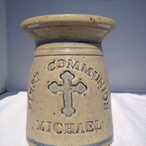 Puede incluir: Un jarrón de cerámica beige con las palabras "FIRST COMMUNION" y "MICHAEL" inscritas alrededor de una cruz. El jarrón tiene una franja azul cerca de la base y una parte superior acampanada.