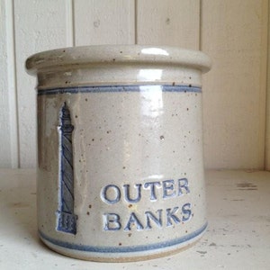 Op de afbeelding: Een witte keramische pot met blauwe rand en een vuurtorenontwerp. De pot heeft de tekst "Outer Banks" in blauwe letters.