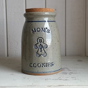 Könnte beinhalten: Eine graue Keramik-Keksdose mit einem Korkdeckel. Die Dose hat ein blaues und weißes Lebkuchenmann-Design und die Worte "Mom's Cookies" auf der Vorderseite.