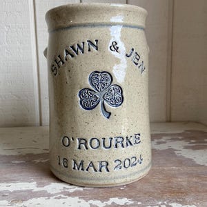 May include: A cream-coloured ceramic jar with a blue shamrock design and the text "Shawn & Jen O'Rourke 16 Mar 2024".