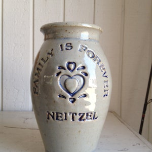 May include: A white ceramic vase with blue lettering that reads "Family is Forever" and a blue heart design. The vase is signed "Neitzel".