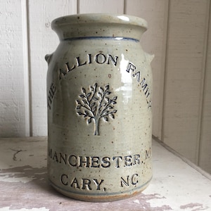 May include: A stoneware jar with a light green glaze. The jar features the text "THE ALLION FAMILY" and "MANCHESTER, MA CARY, NC" in dark blue lettering. A tree design is centered on the jar.
