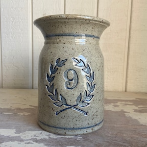May include: A stoneware vase with a speckled glaze in shades of beige and brown. The vase has a blue and white design of a laurel wreath with the number 9 in the centre.