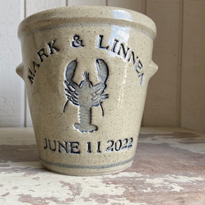 May include: A beige ceramic crock with blue lettering and a lobster design. The crock is inscribed with "Mark & Linnea June 11 2022".