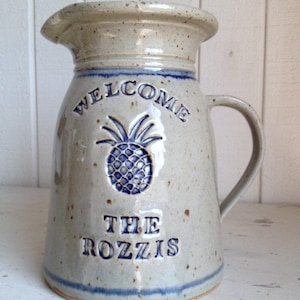 May include: A speckled stoneware pitcher with a wide spout and handle. The pitcher features the word "WELCOME" above a blue pineapple design and the words "THE ROZZIS" below. A blue band accents the base and rim.