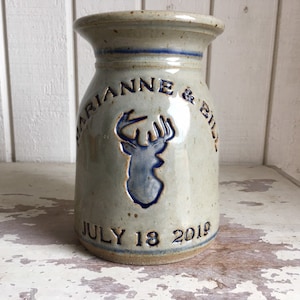 May include: A light gray ceramic vase with a blue glaze. It has a deer silhouette with antlers and the text "Marianne & Bill" and "July 18 2019" on the front.