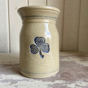 May include: A cream-coloured ceramic vase with a blue and white shamrock design. The vase has a rounded body and a slightly flared opening.