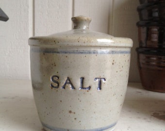 Peut inclure: Un pot à sel en céramique avec un couvercle, présentant un glaçage moucheté dans des tons de gris et de brun. Le pot est doté d'une bande bleue autour du haut et du bas, et le mot "SEL" est inscrit en bleu sur le devant.