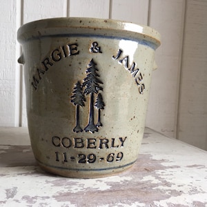 May include: A beige ceramic crock with blue lettering. The crock is decorated with a design of three stylised trees. The text reads "Margie & James Coberly 11-29-69".