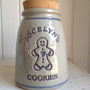 Könnte beinhalten: Eine graue Keramik-Keksdose mit einem Korkdeckel. Die Dose hat ein blaues und weißes Design mit einem Lebkuchenmann und dem Text "Jocelyn's Cookies".