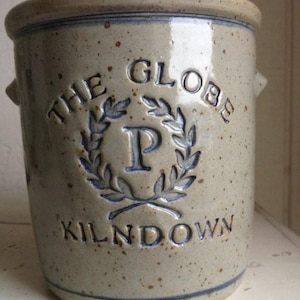 May include: A stoneware crock with a speckled, light grey finish and blue accents. The text "THE GLOBE KILNDOWN" is embossed in blue, with a blue "P" inside a laurel wreath. Features two small side handles.