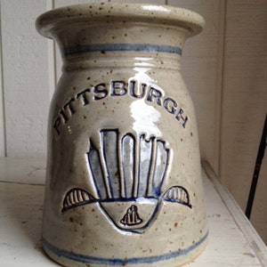 May include: A beige ceramic jar with the word "PITTSBURGH" in blue lettering. The jar features a blue skyline design and two blue stripes. The jar is a decorative item.