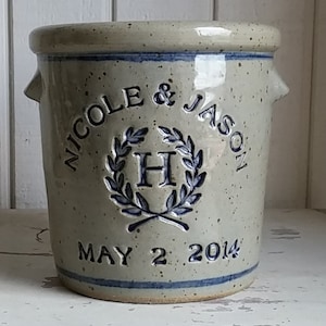 May include: A grey stoneware crock with a blue glaze and a white inscription. The inscription reads "Nicole & Jason" with a wreath and the letter "H" in the centre. The date "May 2 2014" is inscribed below the wreath.