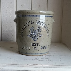 May include: A beige ceramic crock with blue trim and a rooster design. The crock is labelled "Molly's Kitchen Est. Aug 13 2011".