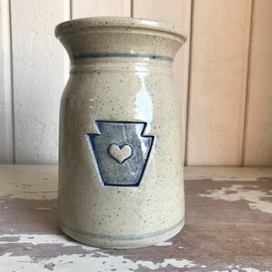 Puede incluir: Un jarrón de cerámica beige moteado con un emblema azul con un corazón. El jarrón tiene una parte superior acampanada y una banda azul alrededor del cuello. Es un artículo de cocina decorativo.