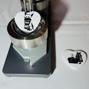 Peut inclure: Une machine à fabriquer des boutons avec un bouton en forme de cœur et un dessin de cheval en noir et blanc. Un autre bouton en forme de cœur avec un cheval et un cavalier est à côté de la machine. La machine est argentée et noire.