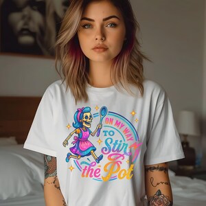 Peut inclure: T-shirt blanc avec un motif coloré d'un chef squelette de dessin animé tenant une cuillère et courant. Le texte "On My Way To Stir the Pot" est affiché en dégradé arc-en-ciel. Un vêtement décontracté pour tous les jours.