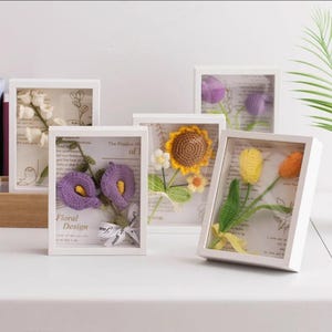 Puede incluir: Una colección de diseños florales enmarcados. Cada marco blanco contiene un arreglo floral de ganchillo, incluyendo calas, tulipanes y un girasol. Los marcos se exhiben sobre una superficie blanca, con el texto "Floral Design" visible en uno.