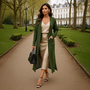 May include: A woman wearing a long, olive green cardigan with pockets, over a cream-colored satin dress. She is carrying a black handbag and wearing black high-heeled sandals. The background shows a park with trees and a building.