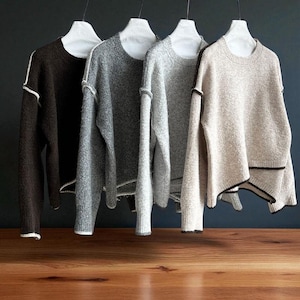K&ouml;nnte beinhalten: Vier Rundhals-Pullover in Braun-, Grau- und Beiget&ouml;nen h&auml;ngen an wei&szlig;en Kleiderb&uuml;geln. Jeder Pullover hat ein strukturiertes Strickmuster und kontrastierende Paspeln am Ausschnitt, an den Manschetten und am Saum. Die Pullover werden vor einem dunklen Hintergrund pr&auml;sentiert.