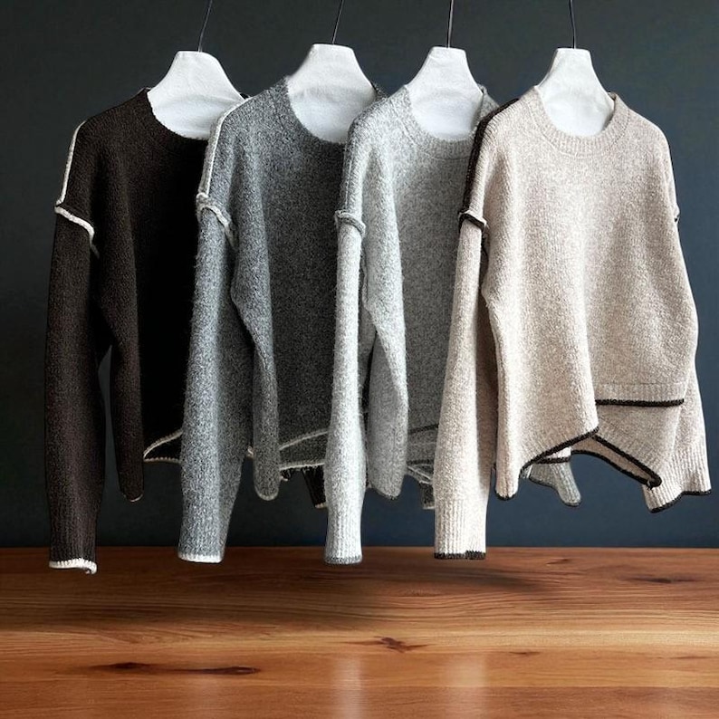 K&ouml;nnte beinhalten: Vier Rundhals-Pullover in Braun-, Grau- und Beiget&ouml;nen h&auml;ngen an wei&szlig;en Kleiderb&uuml;geln. Jeder Pullover hat ein strukturiertes Strickmuster und kontrastierende Paspeln am Ausschnitt, an den Manschetten und am Saum. Die Pullover werden vor einem dunklen Hintergrund pr&auml;sentiert.