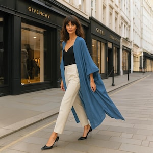 Puede incluir: Una mujer con una chaqueta larga y fluida de estilo kimono azul, una blusa negra y pantalones color crema. Lleva zapatos de tacón negros. El fondo muestra tiendas con los nombres Givenchy y Gucci.