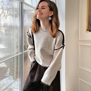 K&ouml;nnte beinhalten: Ein hellgrauer Pullover mit schwarzen Verzierungen an Schultern und &Auml;rmeln. Der Pullover wird mit einer dunkelbraunen Hose kombiniert. Die Person tr&auml;gt silberne Ohrringe. Der Hintergrund umfasst ein Fenster und eine wei&szlig;e T&uuml;r.