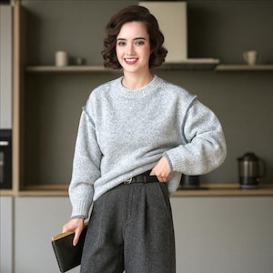 K&ouml;nnte beinhalten: Eine Frau tr&auml;gt einen hellgrauen Pullover mit dunkelgrauen N&auml;hten und eine dunkelgraue Hose. Der Pullover hat einen Rundhalsausschnitt und lange &Auml;rmel. Sie h&auml;lt eine schwarze Clutch und tr&auml;gt einen schwarzen G&uuml;rtel. Der Hintergrund ist eine K&uuml;che.