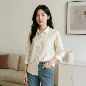 May include: A cream-coloured button-down shirt with a delicate floral pattern in yellow and green. The shirt has long sleeves and is paired with blue jeans. The overall style is casual and feminine.