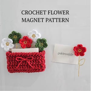 Op de afbeelding: Een rode gehaakte mand met een strik, met witte, rode en groene gehaakte bloemen met gouden centra. De tekst "CROCHET FLOWER MAGNET PATTERN" staat bovenaan. Rechts staat een enkele rode bloem met een gouden steel.
