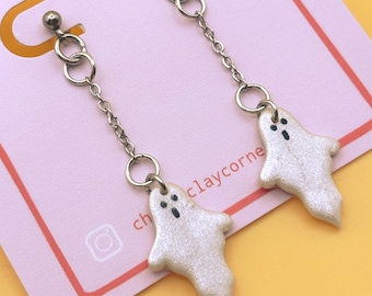 May include: A pair of dangle earrings featuring white, shimmering ghost charms suspended from silver-toned metal chains and stud posts. The earrings are attached to a light pink display card printed with the text CHLOESCLAY CORNER in colorful letters.