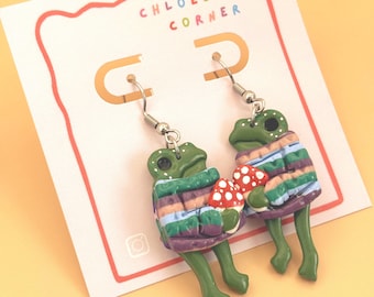 May include: A pair of dangle earrings featuring green frogs wearing striped sweaters in shades of purple, blue, and green, each holding a red and white spotted mushroom. The earrings are displayed on a white card labeled CHLOESCLAY CORNER.