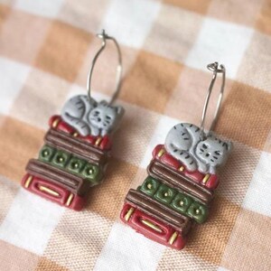 May include: A pair of silver hoop earrings. Each earring features a gray cat sleeping on a stack of books. The books are red, green, and brown with gold accents. The earrings are set against a checkered background.
