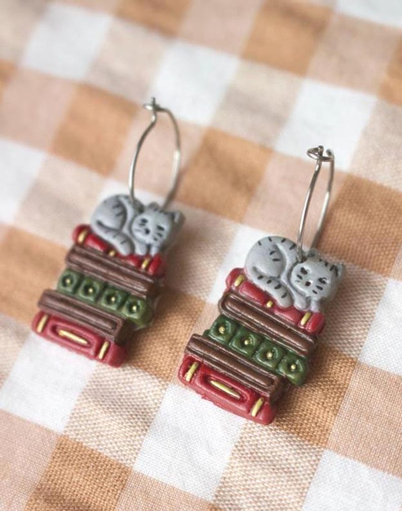 May include: A pair of silver hoop earrings. Each earring features a gray cat sleeping on a stack of books. The books are red, green, and brown with gold accents. The earrings are set against a checkered background.
