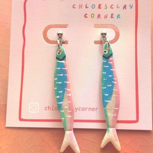 K&ouml;nnte beinhalten: Ein Paar h&auml;ngende Ohrringe in Fischform mit einem bunten Design aus blauen, gr&uuml;nen und rosafarbenen Segmenten, wei&szlig;en Akzenten und einer glitzernden Oberfl&auml;che. Die Ohrringe sind auf einer wei&szlig;en Karte mit der Aufschrift CHLOESCLAY CORNER befestigt.