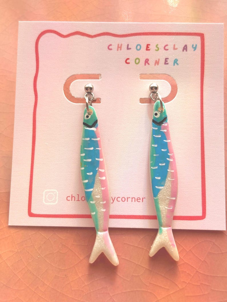 K&ouml;nnte beinhalten: Ein Paar h&auml;ngende Ohrringe in Fischform mit einem bunten Design aus blauen, gr&uuml;nen und rosafarbenen Segmenten, wei&szlig;en Akzenten und einer glitzernden Oberfl&auml;che. Die Ohrringe sind auf einer wei&szlig;en Karte mit der Aufschrift CHLOESCLAY CORNER befestigt.