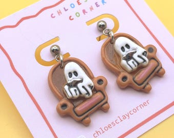 May include: A pair of dangle earrings featuring a white ghost sitting in a brown armchair. The earrings are attached to a light pink display card with the text CHLOESCLAY CORNER and chloesclaycorner printed in black and colorful letters.