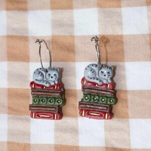 May include: A pair of earrings featuring a gray cat curled up on a stack of books. The books are red, green, and brown with gold accents. The earrings have silver-colored hoops.