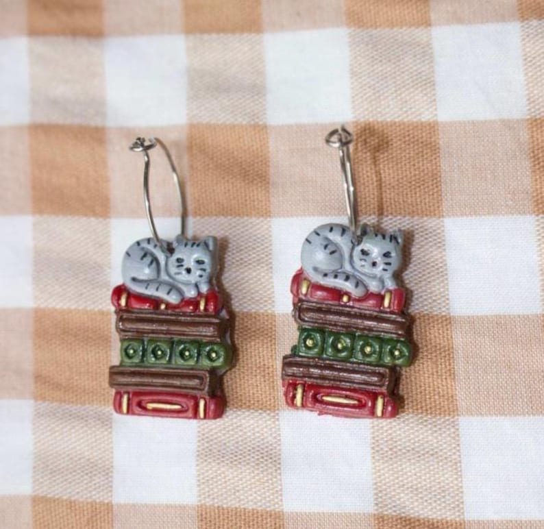 May include: A pair of earrings featuring a gray cat curled up on a stack of books. The books are red, green, and brown with gold accents. The earrings have silver-colored hoops.