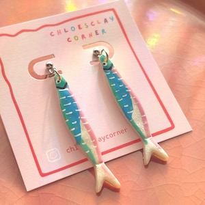K&ouml;nnte beinhalten: Ein Paar Ohrh&auml;nger in Form von Sardinen mit blau, gr&uuml;n und rosa bemalten Details und einem schimmernden Finish. Die Ohrringe sind an einer wei&szlig;en Pr&auml;sentationskarte befestigt, die mit dem Text CHLOESCLAY CORNER und einem Instagram-Symbol bedruckt ist.
