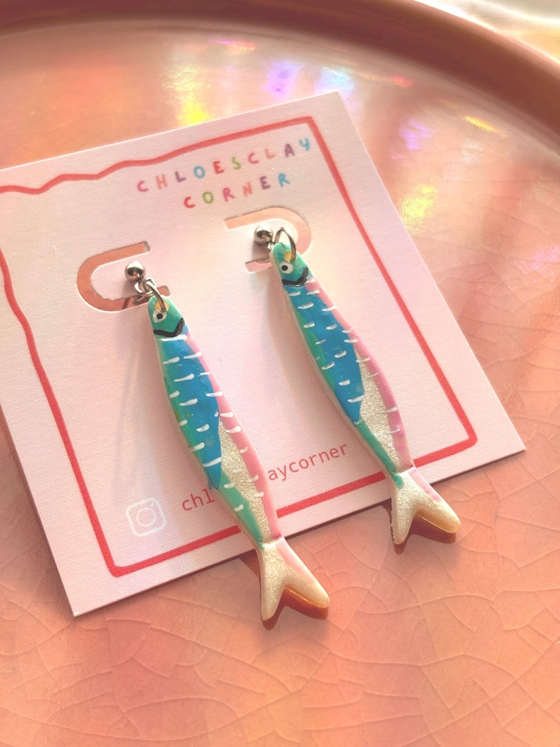 K&ouml;nnte beinhalten: Ein Paar Ohrh&auml;nger in Form von Sardinen mit blau, gr&uuml;n und rosa bemalten Details und einem schimmernden Finish. Die Ohrringe sind an einer wei&szlig;en Pr&auml;sentationskarte befestigt, die mit dem Text CHLOESCLAY CORNER und einem Instagram-Symbol bedruckt ist.