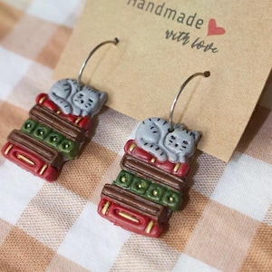 May include: A pair of handmade earrings featuring a gray cat sleeping on a stack of books. The books are red, green, and brown. The earrings are attached to silver hoops and displayed on a tan card with the text "Handmade with love".