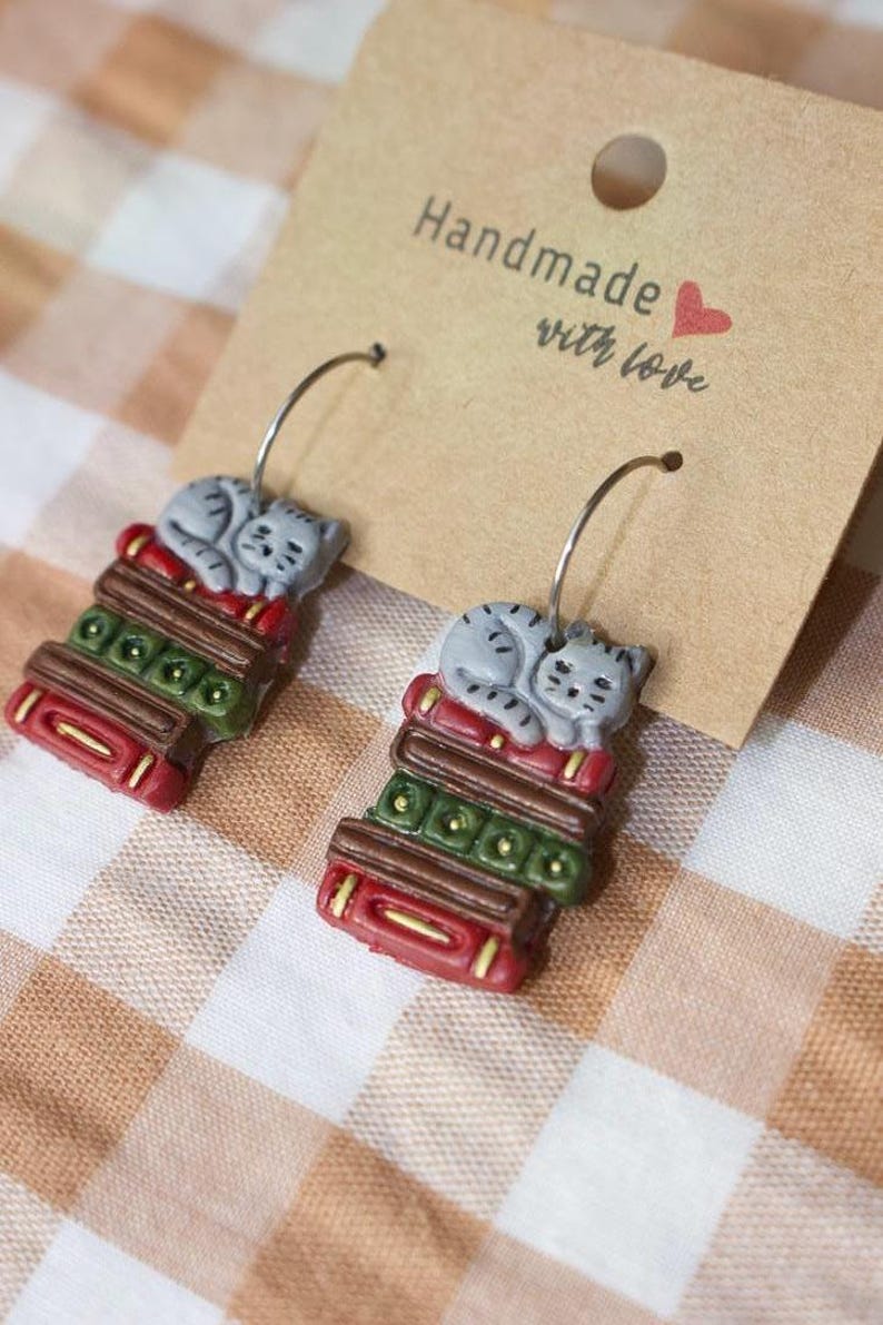 May include: A pair of handmade earrings featuring a gray cat sleeping on a stack of books. The books are red, green, and brown. The earrings are attached to silver hoops and displayed on a tan card with the text "Handmade with love".
