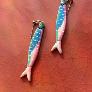 K&ouml;nnte beinhalten: Ein Paar fischf&ouml;rmige Ohrh&auml;nger in einer lebendigen Farbpalette aus Blau, Pink und Gr&uuml;n. Jeder Ohrring verf&uuml;gt &uuml;ber einen silberfarbenen Stecker und eine texturierte, glitzernde Oberfl&auml;che am Schwanzteil mit aufgemalten Schuppendetails.