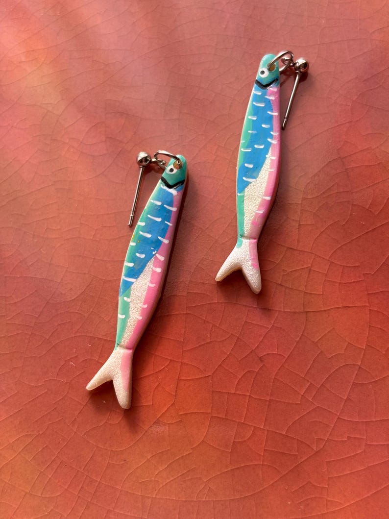 K&ouml;nnte beinhalten: Ein Paar fischf&ouml;rmige Ohrh&auml;nger in einer lebendigen Farbpalette aus Blau, Pink und Gr&uuml;n. Jeder Ohrring verf&uuml;gt &uuml;ber einen silberfarbenen Stecker und eine texturierte, glitzernde Oberfl&auml;che am Schwanzteil mit aufgemalten Schuppendetails.