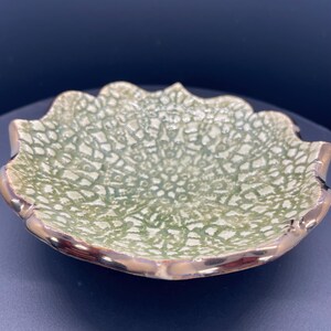 May include: A decorative, ceramic bowl with a scalloped edge. The bowl has a textured, light green interior with a gold-colored rim. The bowl's design is reminiscent of a flower or leaf, making it a unique piece for home decor.