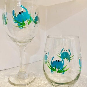 May include: Two clear wine glasses, one stemmed and one stemless, decorated with hand-painted blue crabs and green seaweed. The crab designs have black eyes. The glasses are on a white surface.